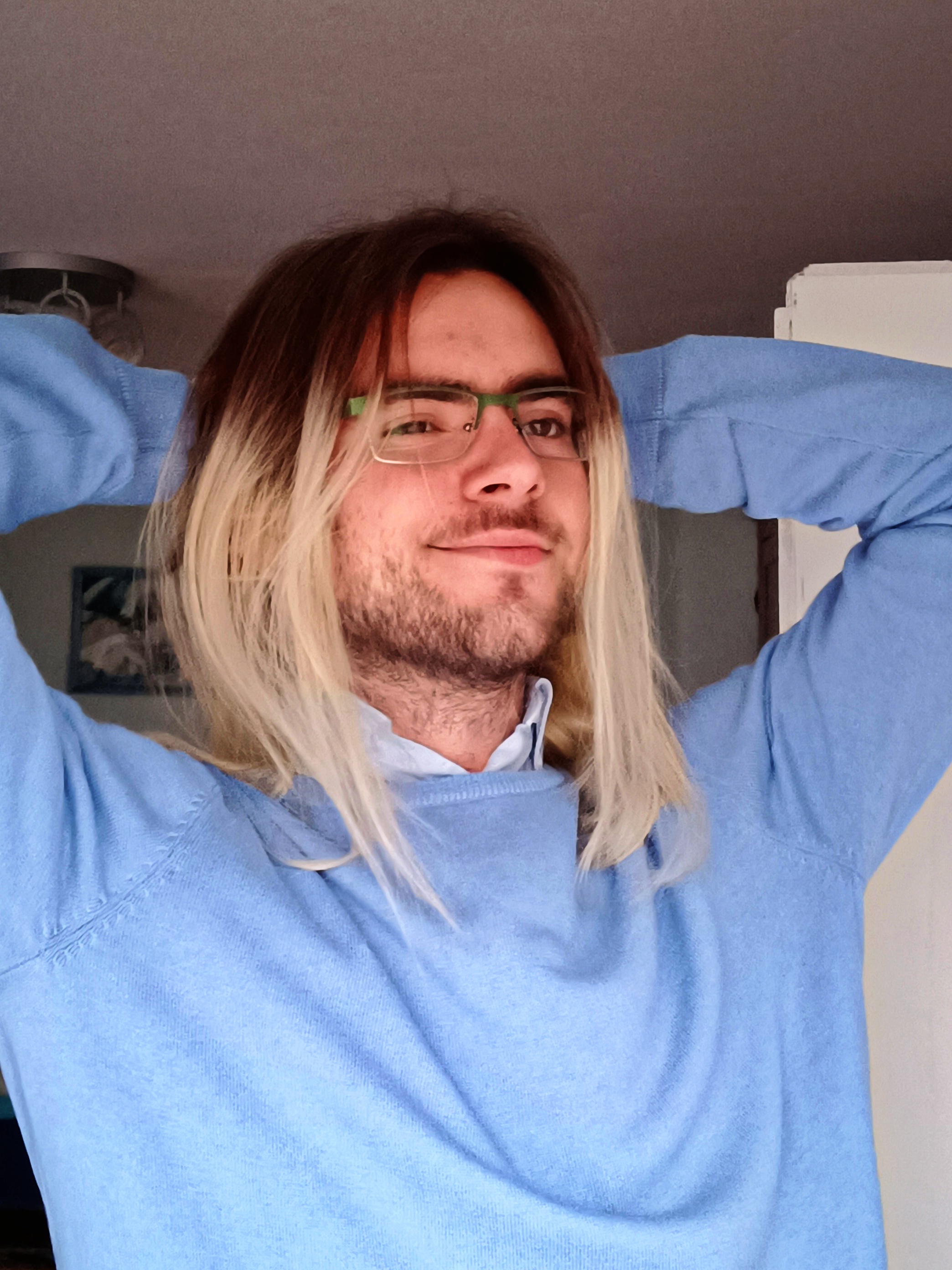 Dmitry with shoulder-length brown-to-blonde ombre hair, green-framed glasses, and a light blue sweater, posing with hands behind head and a slight smile. Background: room with white door and ceiling light.