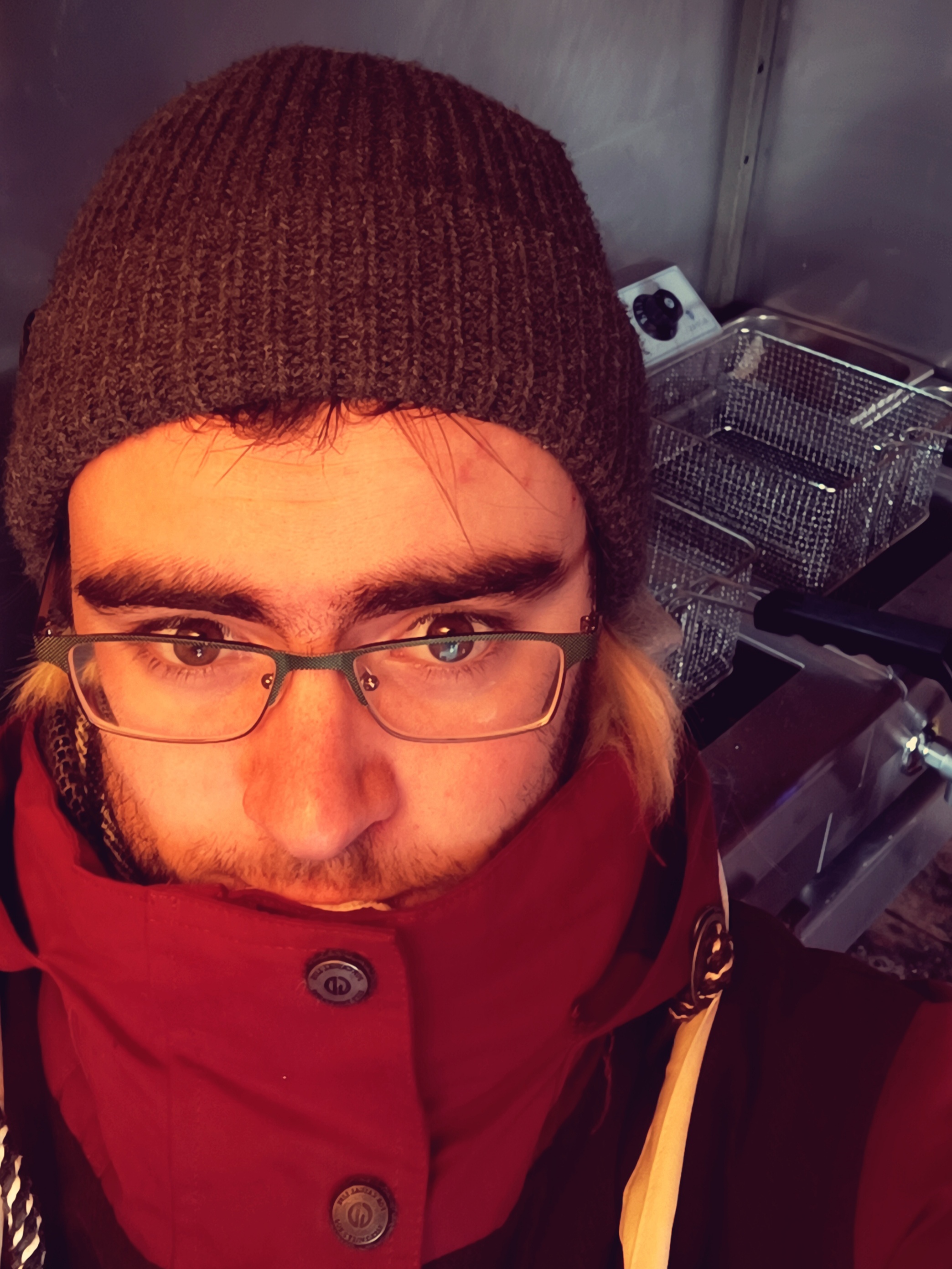 Dmitry in a red winter coat with the collar up, dark knitted beanie, and green-framed glasses. Serious expression. Background: stainless steel kitchen with deep fryers.