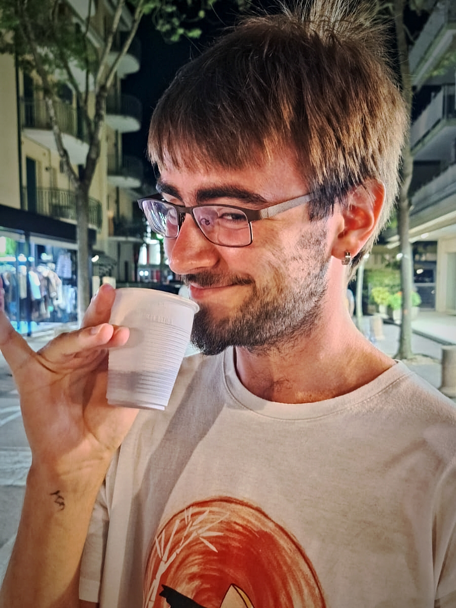 Dmitry mit kurzem braunem Haar, einem stoppeligem Kinnbart, braun gerahmter Brille und einem weißen T-Shirt, hält einen Plastik-Becher am Mund und grinst in die Kamera. Hintergrund: Nächtliche beleuchtete Straße.