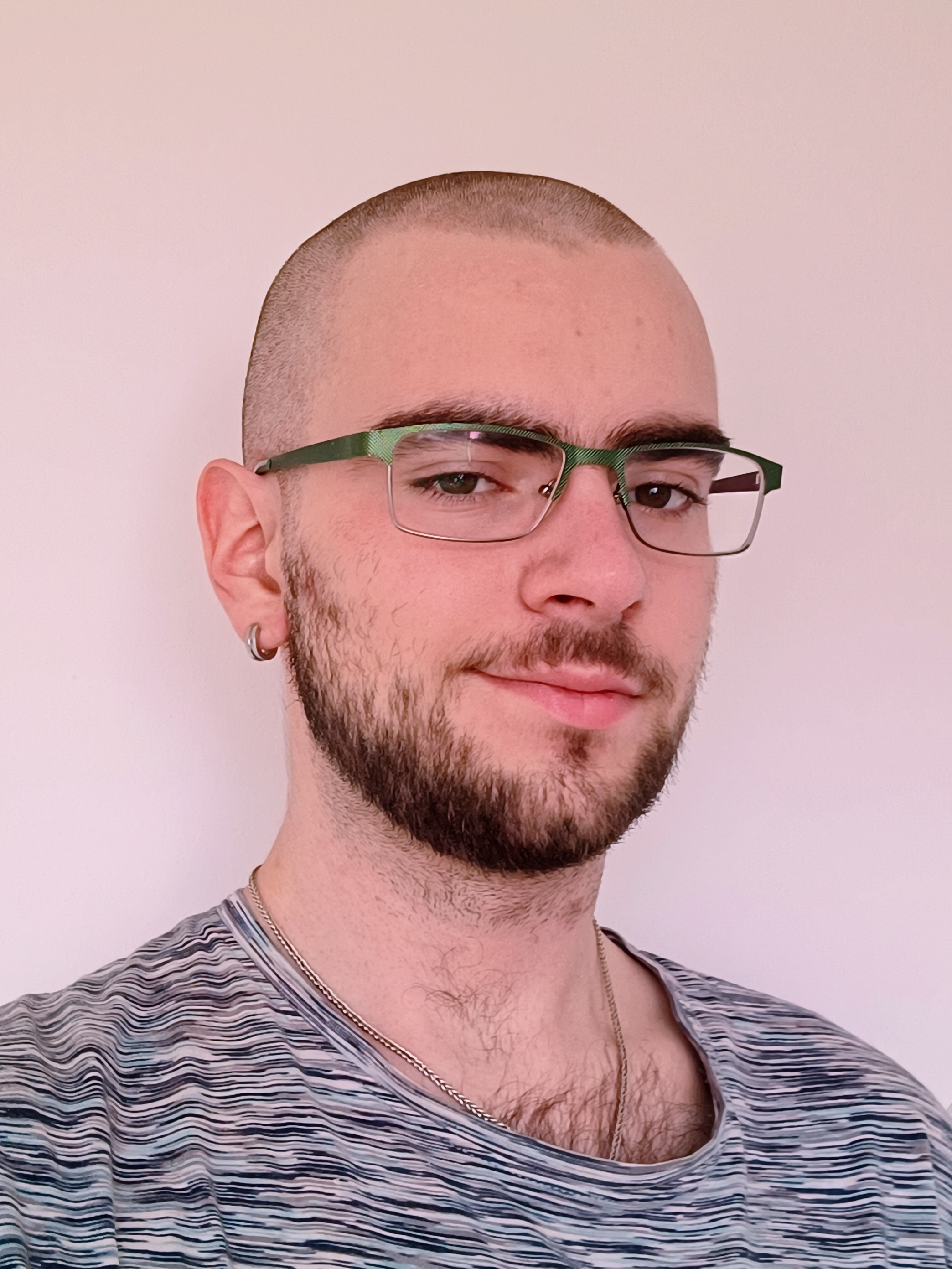 Dmitry with a shaved head, trimmed beard, green-framed glasses, and a striped t-shirt, smiling subtly. Wearing a silver necklace. Background: light-colored wall.
